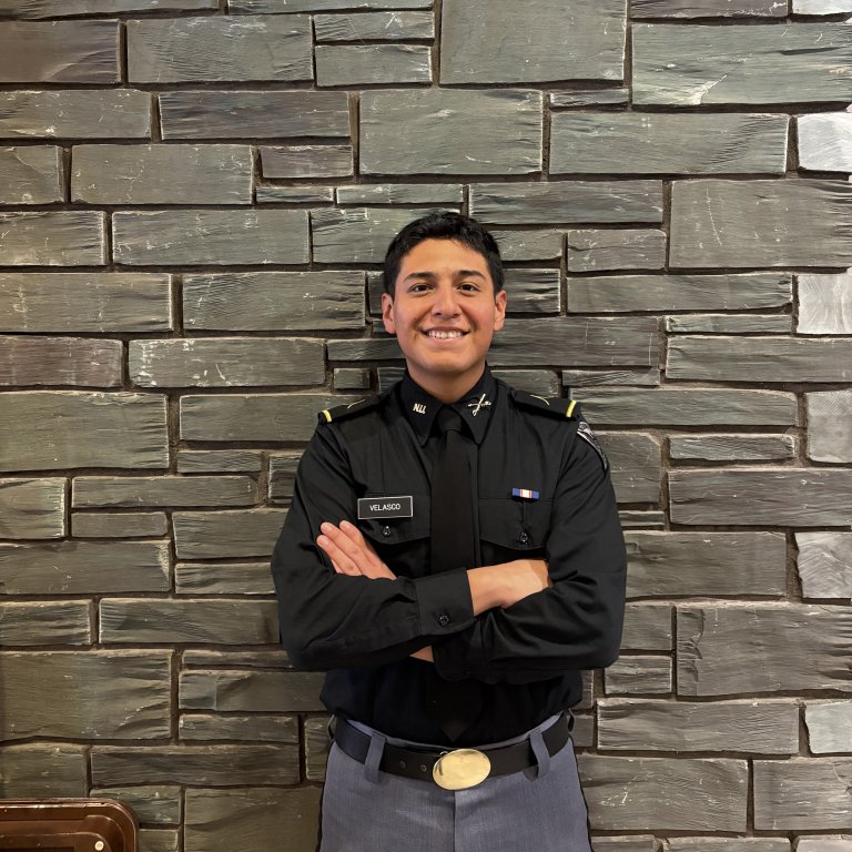 Person in a dark uniform stands with arms crossed in front of a stone brick wall.