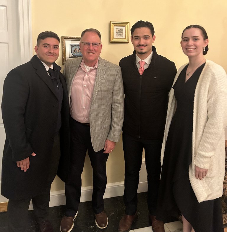 Four people stand together indoors in formal and semi‑formal attire near a light‑colored wall with framed pictures.