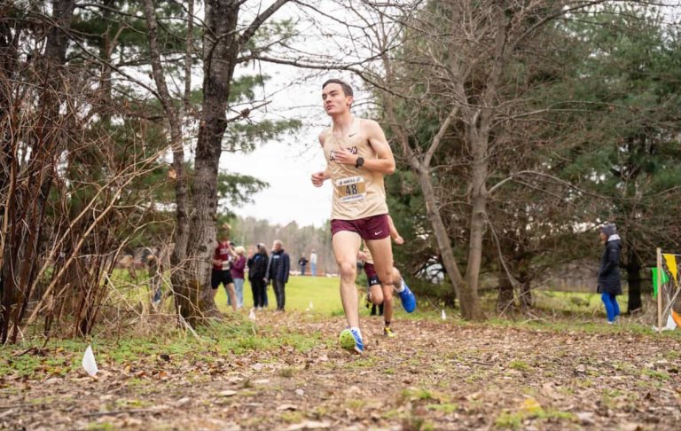 A runner in athletic gear races along a leaf‑covered trail with trees and spectators in the background.