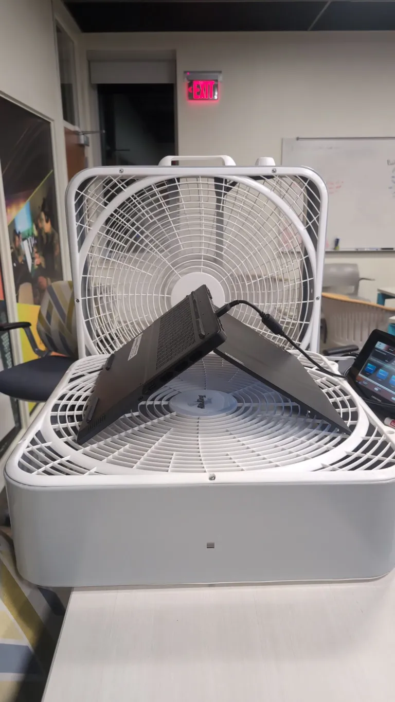 A laptop sits open on its side on top of a large box fan with another fan placed behind it in an office setting.