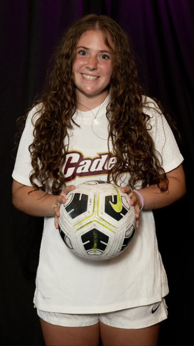 Person in a white athletic uniform holds a soccer ball against a dark background.