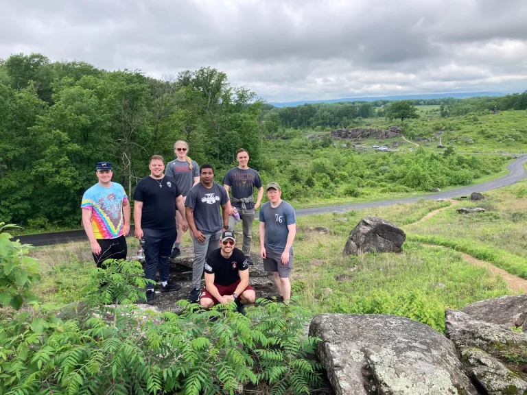 Seven people stand on a rocky hill with green fields, trees, and a winding road in the background.