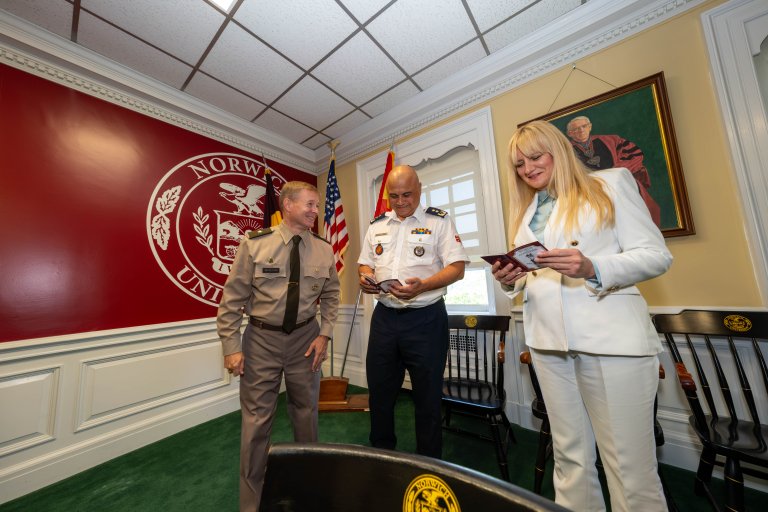 Three people in formal uniforms and a light suit stand in a room with Norwich University signage, flags, and framed art.