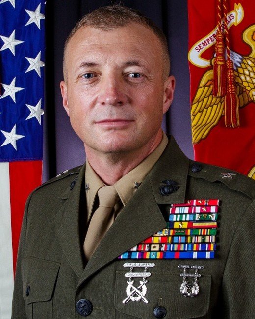 Person in a decorated military uniform stands before U.S. and Marine Corps flags.