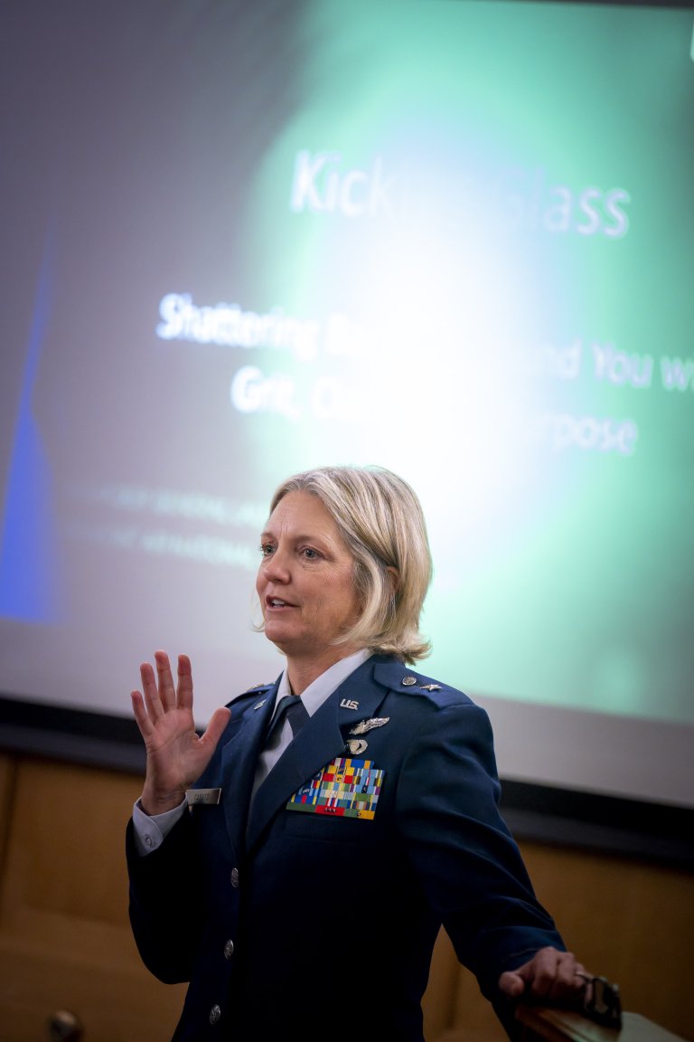Alt Text: A person in a military dress uniform gestures while standing in front of a projected slide with the title “Kicking Glass.”