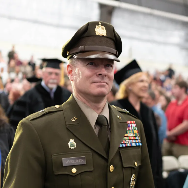 Person in a decorated military uniform stands indoors at a ceremony with people seated and standing in the background.