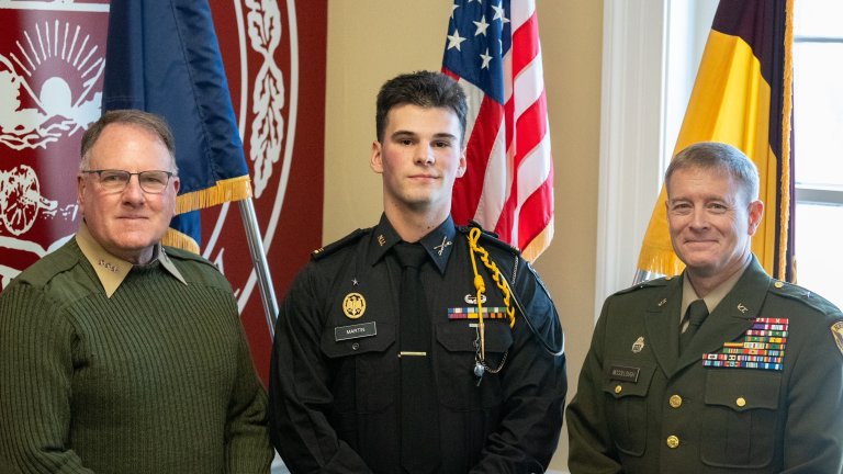Three people in uniform stand in front of a large university seal and two flags.
