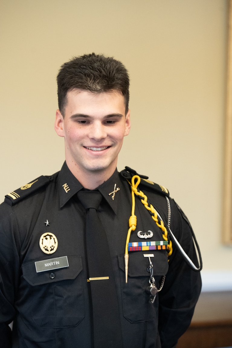 A person in a dark military-style uniform with shoulder cords, badges, and ribbons stands indoors against a plain wall.
