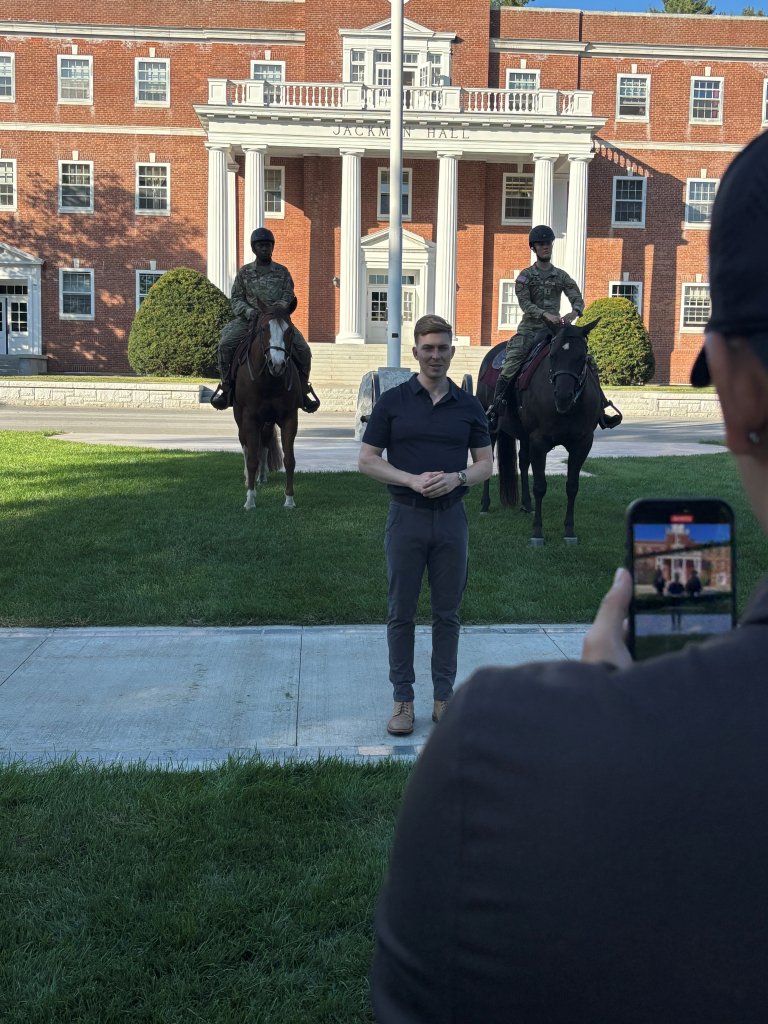 A person stands on a sidewalk while two riders on horses face forward in front of a large brick building with white columns.