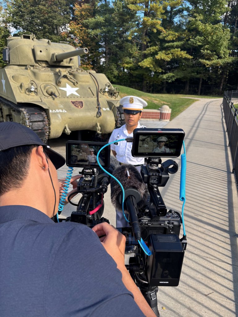 A person in a white uniform stands in front of a military tank while someone operates two video cameras aimed at them.