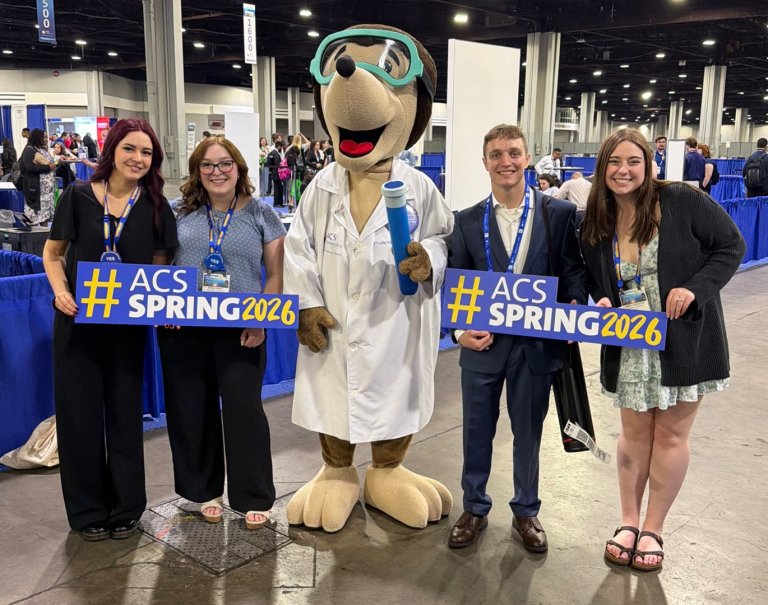 Four attendees pose with an ACS mascot and ACS Spring 2026 signs in a convention hall.