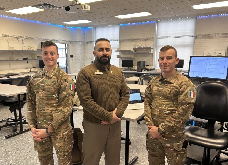 Two cadets and an instructor stand in a computer lab classroom.