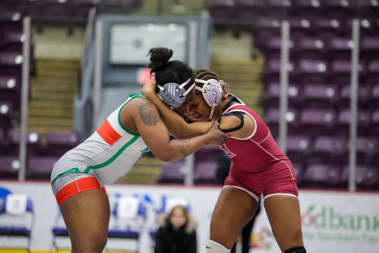 Two wrestlers grip each other in a neutral position on a mat inside an arena with rows of purple seats.