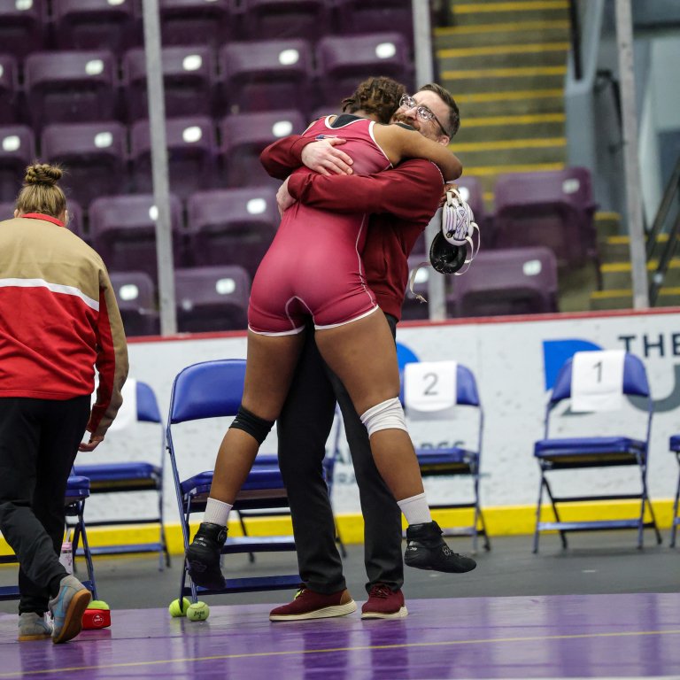 A person in a maroon wrestling uniform hugs another person while lifting off the mat near blue chairs in an arena.