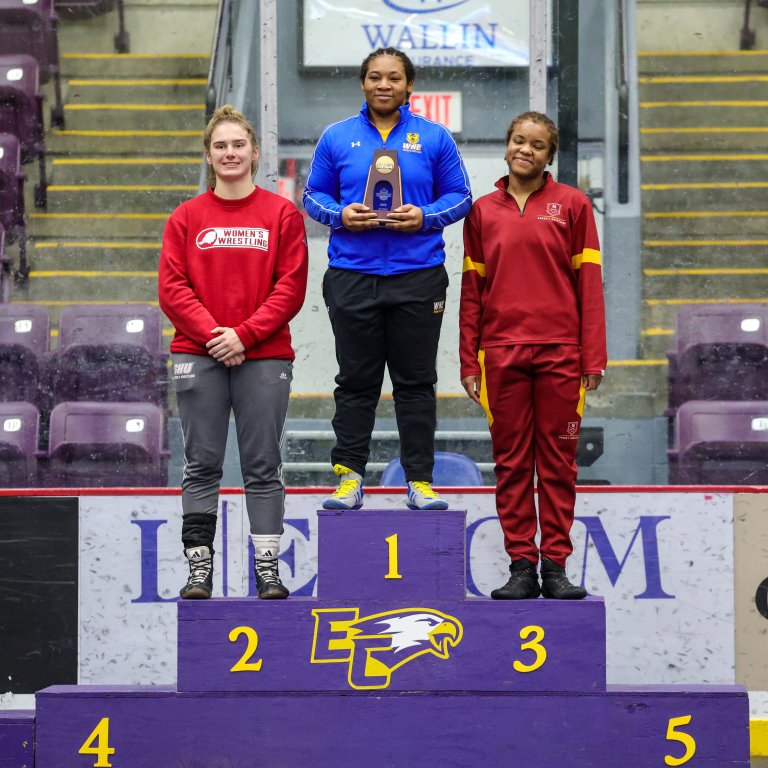 Three athletes stand on a purple podium with numbered steps, with the center athlete holding a small award.