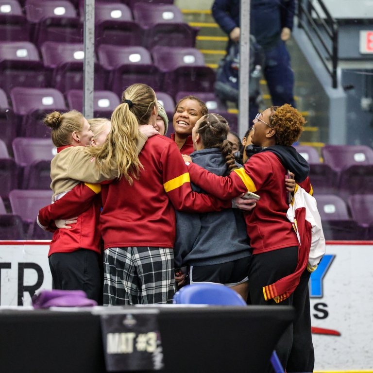A group of people in red and gray team gear stand in a tight huddle near purple arena seats.