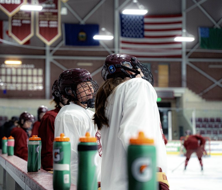 Hockey players in white and maroon jerseys stand by the bench during practice on an indoor ice rink.