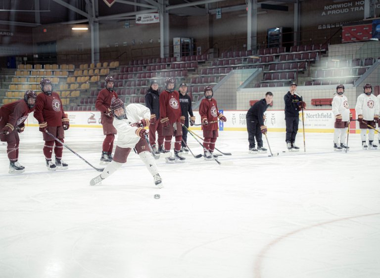 Hockey player in a white jersey shoots the puck as teammates and coaches watch on the ice.