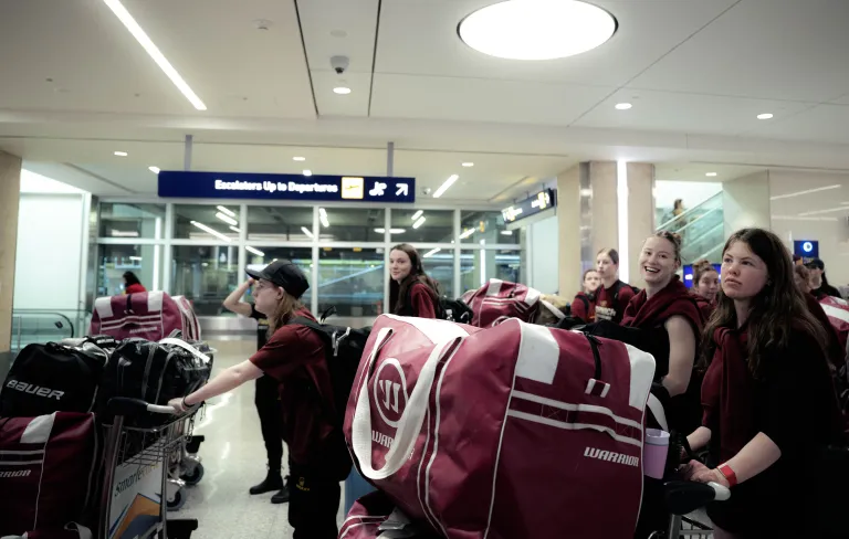 Students with luggage carts loaded with duffel bags wait near the departures escalator in an airport terminal.