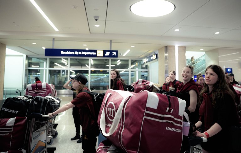 Students with luggage carts loaded with duffel bags wait near the departures escalator in an airport terminal.