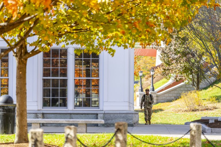 A person in camouflage walks on a campus path near a white building with large windows and fall trees.