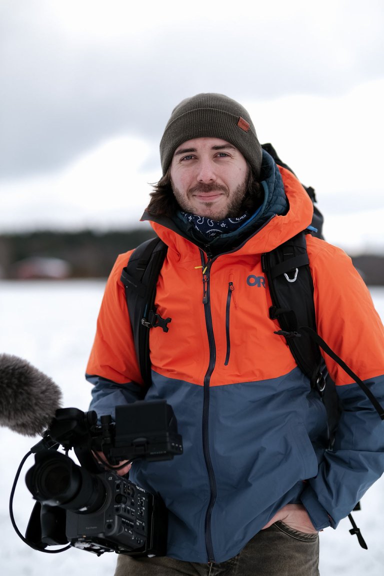 Person in an orange and blue jacket holds a large video camera while standing outdoors in a snowy setting.