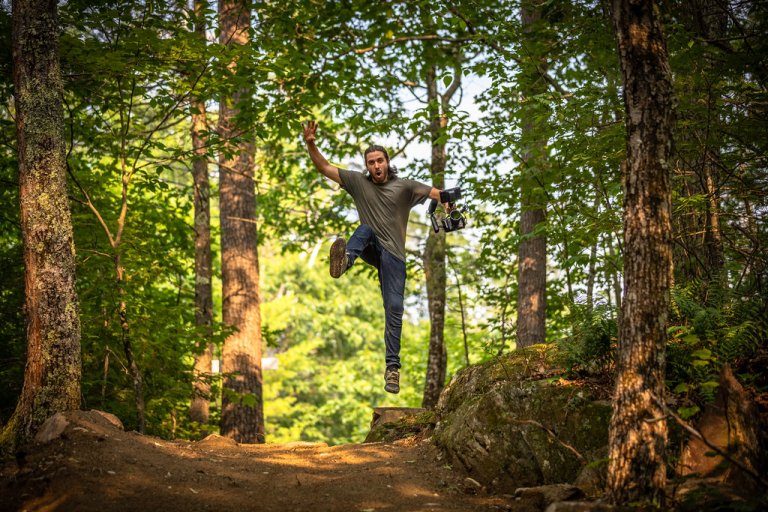 A person jumps mid‑air on a forest trail holding a camera with sunlight filtering through the trees.
