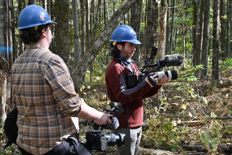Two people wearing hard hats stand in a forest as one holds a video camera with a large microphone.