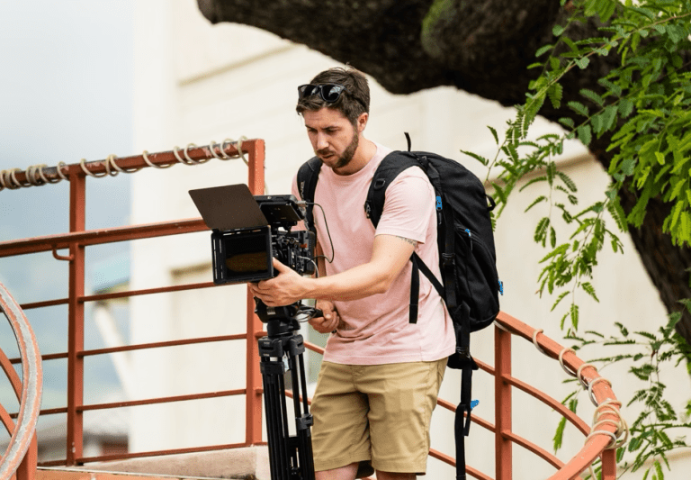 Person with a backpack adjusts a video camera on a tripod while standing on an outdoor staircase near a tree.