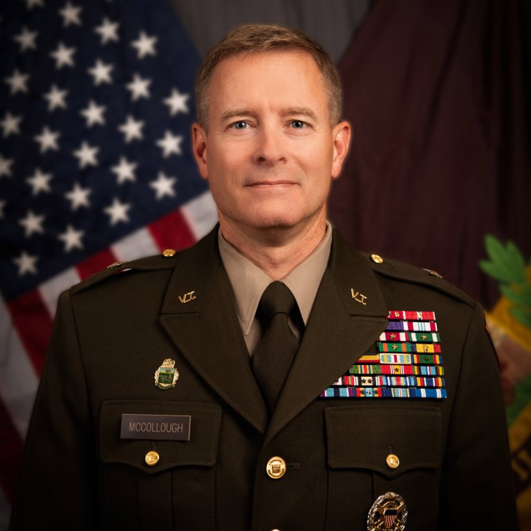 Uniformed military officer stands in front of flags.