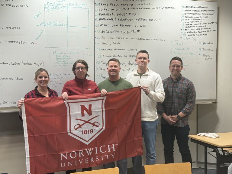 Five people stand in a classroom holding a red Norwich University flag in front of large whiteboards filled with handwritten notes.