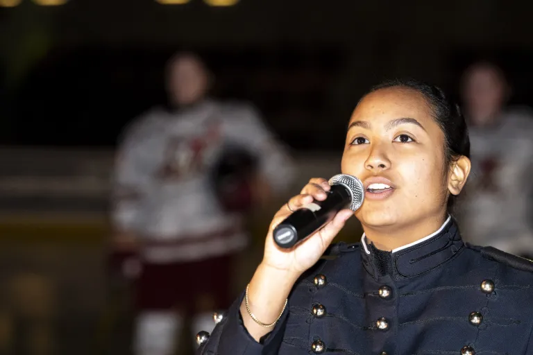 Cadet in dress uniform sings into a microphone indoors.