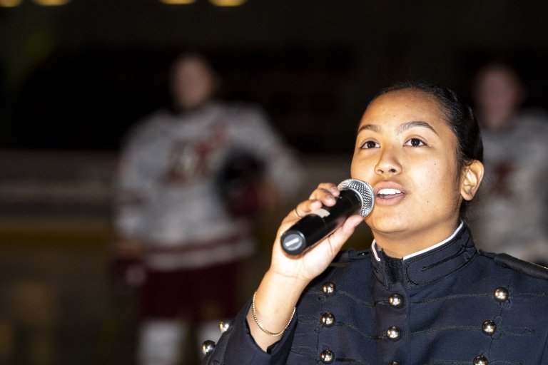 Cadet in dress uniform sings into a microphone indoors.
