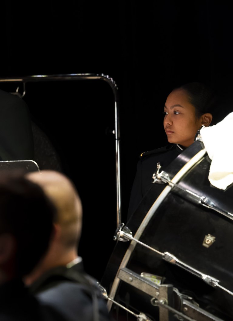 Cadet in dress uniform stands beside a large drum on a dark stage.