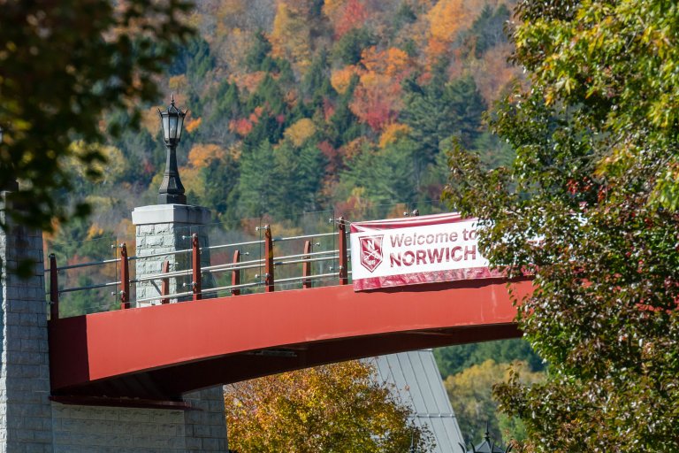 A red bridge with a “Welcome to Norwich” banner spans over trees with colorful fall foliage on a hillside.