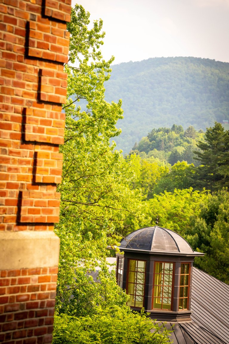 A brick building stands beside green trees with a round rooftop window structure and forested hills in the background.