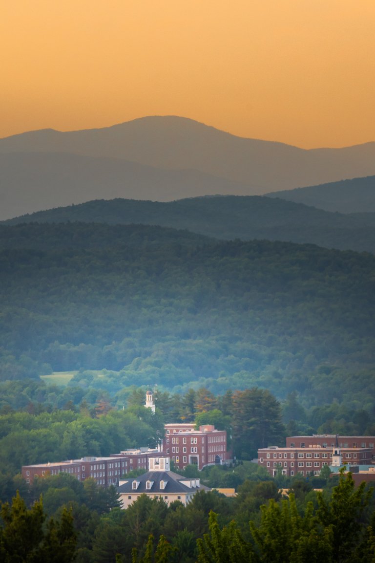 A cluster of red brick campus buildings sits among dense green forests with layered mountains under an orange sky.