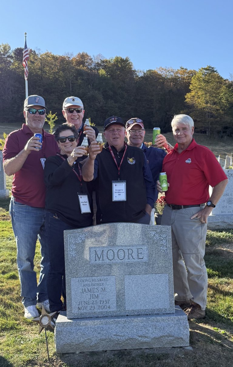 Five people stand behind a Moore headstone in a cemetery holding up drink cans with trees and a flagpole in the background.