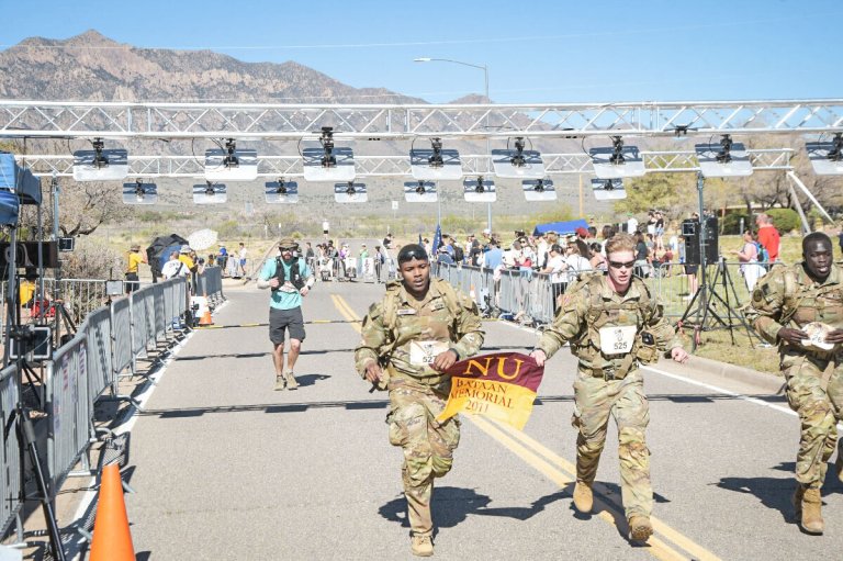 Cadets in uniform cross the finish line carrying an NU Bataan Memorial flag.
