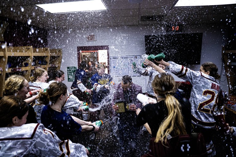 Women's hockey players spray water in a locker room as a person in a suit holds a plaque.