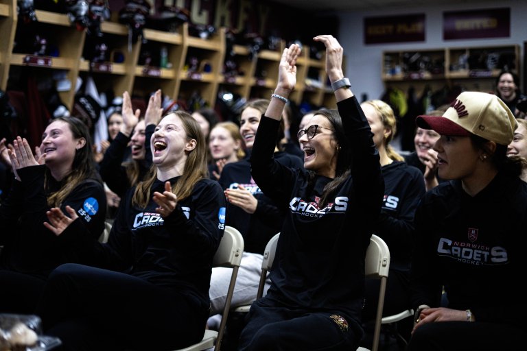 Norwich women's hockey players cheer and clap while seated in a locker room.
