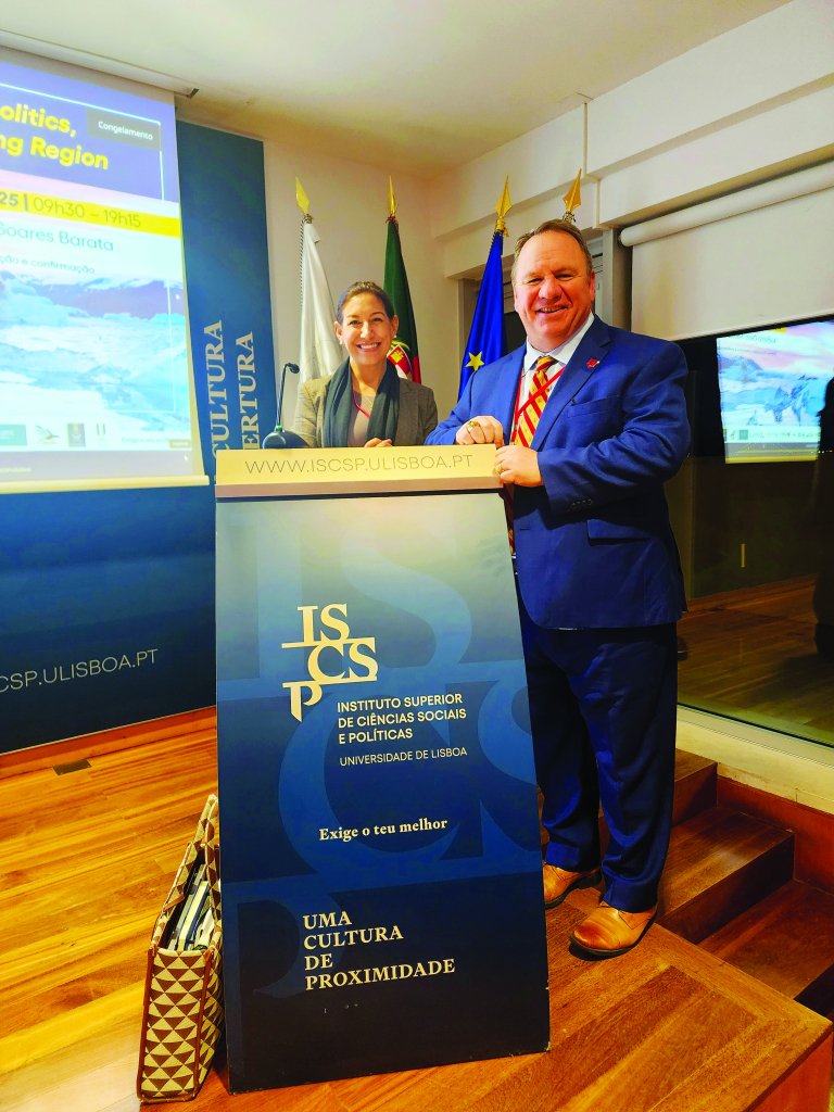 Two speakers stand at an ISCSP podium in a conference room with flags behind them.