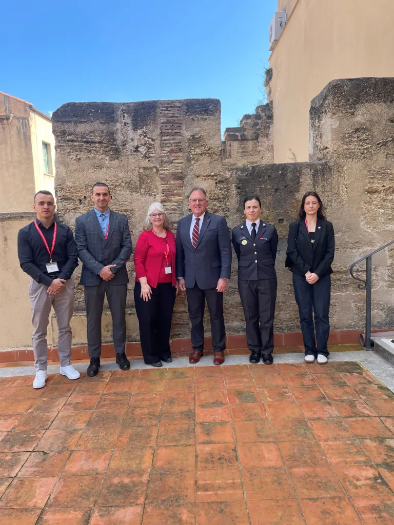 Six people stand on a tiled courtyard in front of a stone wall.