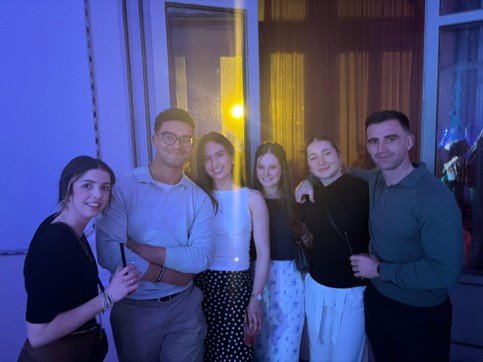 Six people stand close together indoors near large windows.