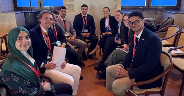 Students in suits sit in a circle of chairs in a room with stone walls.