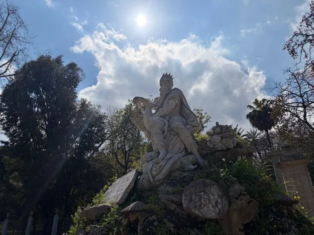Stone statue of seated figures rises under a bright sun and cloudy sky.