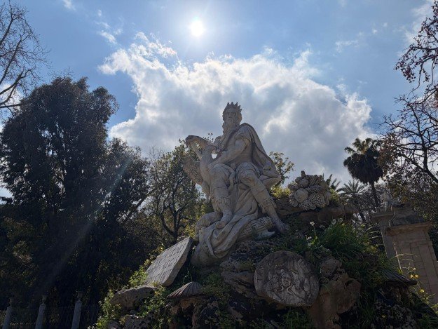Stone statue of seated figures rises under a bright sun and cloudy sky.