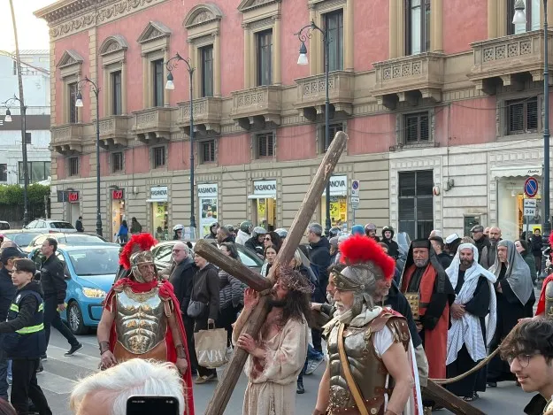 Costumed people walk in a street procession with a person carrying a wooden cross.