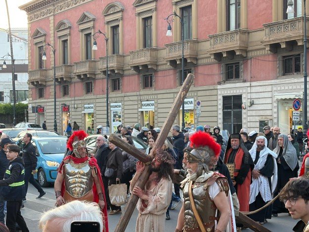 Costumed people walk in a street procession with a person carrying a wooden cross.