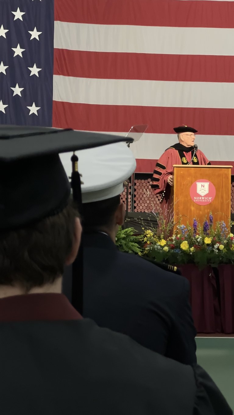 Speaker in academic regalia stands at a Norwich University podium before a large American flag while graduates listen.